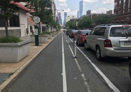 Image from Local Road Safety presentation. Image shows street with wide sidewalks and tree boxes as a landscape buffer between a designate bike line. The bike lane is separated from traffic by white delineators and a row of parked cars. This road design shows that this community prioritizes walking and biking. 