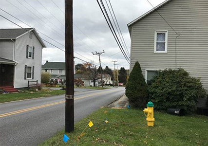 Image from Local Road Safety presentation. Image shows two lane road with double yellow lines. There are no sidewalks or other pedestrian/bicyclist facilities. The houses and telephone poles are very close to the edge of the road. This photo shows the priority on this road is to move cars from Point A to Point B as quickly as possible.